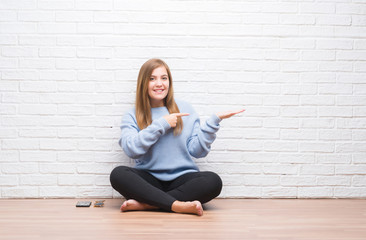 Young adult woman sitting on the floor in autumn over white brick wall amazed and smiling to the camera while presenting with hand and pointing with finger.