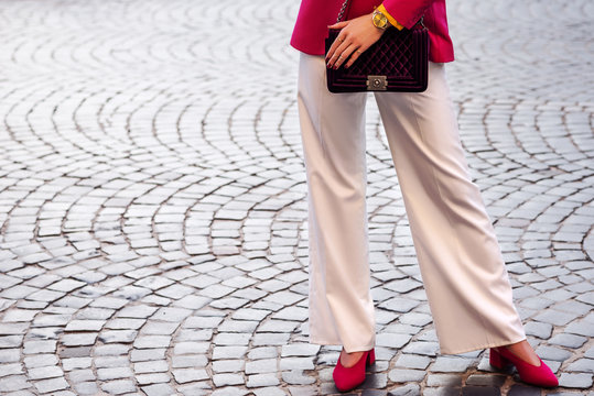 Fashion details: woman wearing white wide-leg trousers, pink blazer, wrist watch, pointed toe shoes, holding velvet violet quilted bag, posing in street of european city. Copy, empty space for text