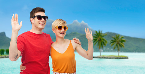 travel, tourism and summer holidays concept - smiling couple in sunglasses hugging and waving hands at touristic resort over bora bora island beach background - obrazy, fototapety, plakaty