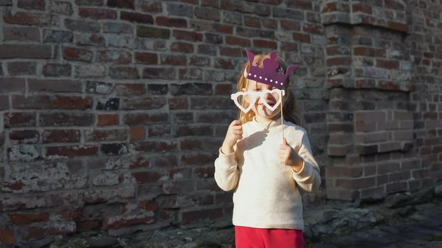 Little cute girl playing with paper phony glasses and a crown on a stick next to a brick wall.