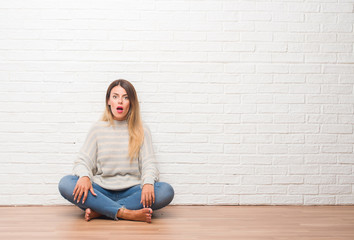 Fototapeta na wymiar Young adult woman sitting on the floor over white brick wall at home afraid and shocked with surprise expression, fear and excited face.