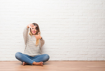 Young adult woman sitting on the floor over white brick wall at home smiling making frame with hands and fingers with happy face. Creativity and photography concept.