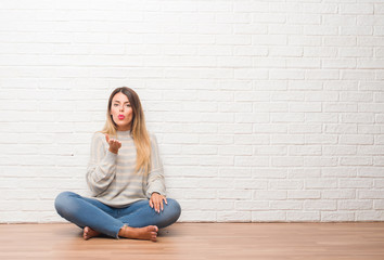 Young adult woman sitting on the floor over white brick wall at home looking at the camera blowing a kiss with hand on air being lovely and sexy. Love expression.