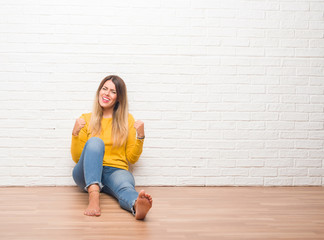 Young adult woman sitting on the floor over white brick wall very happy and excited doing winner gesture with arms raised, smiling and screaming for success. Celebration concept.