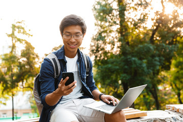 Smiling asian male student in eyeglasses using smartphone
