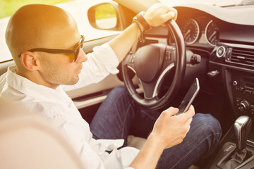 Young man texting and driving. Guy driving a car and using smartphone. Closeup, back light, medium retouch.