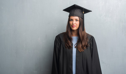 Young brunette woman over grunge grey wall wearing graduate uniform with a confident expression on...