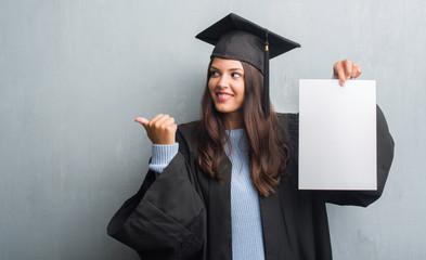 Young brunette woman over grunge grey wall wearing graduate uniform holding degree pointing and showing with thumb up to the side with happy face smiling