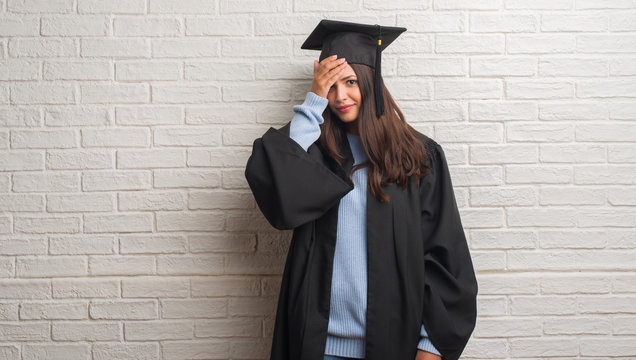 Young brunette woman standing over white brick wall wearing graduate uniform stressed with hand on head, shocked with shame and surprise face, angry and frustrated. Fear and upset for mistake.
