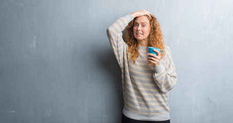 Fototapeta premium Young redhead woman over grey grunge wall drinking a cup of coffee stressed with hand on head, shocked with shame and surprise face, angry and frustrated. Fear and upset for mistake.