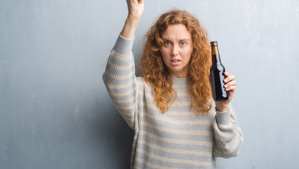 Young redhead woman over grey grunge wall holding beer bottle annoyed and frustrated shouting with anger, crazy and yelling with raised hand, anger concept