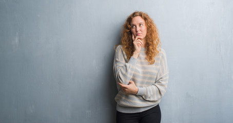 Young redhead woman over grey grunge wall with hand on chin thinking about question, pensive expression. Smiling with thoughtful face. Doubt concept.