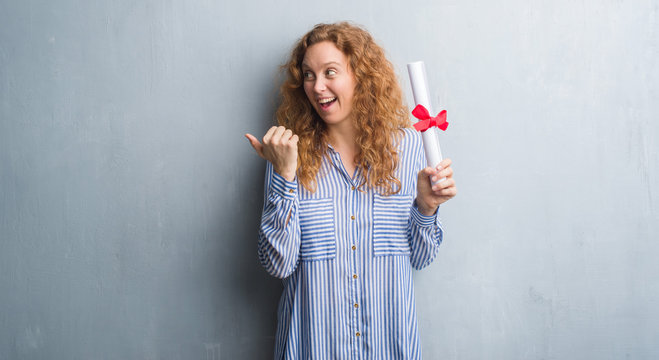 Young redhead business woman over grey grunge wall holding diploma pointing and showing with thumb up to the side with happy face smiling