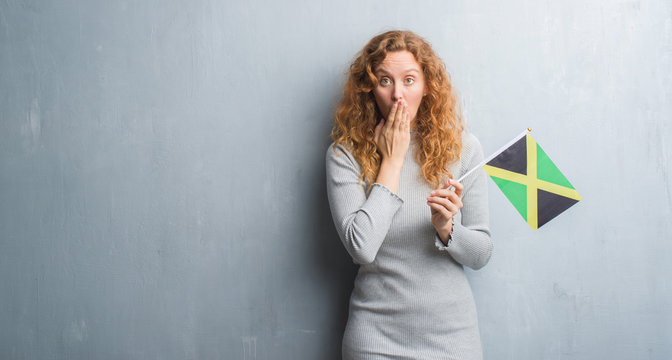 Young redhead woman over grey grunge wall holding flag of Jamaica cover mouth with hand shocked with shame for mistake, expression of fear, scared in silence, secret concept