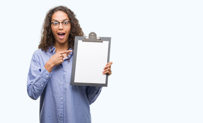 Fototapeta premium Young hispanic business woman holding clipboard very happy pointing with hand and finger