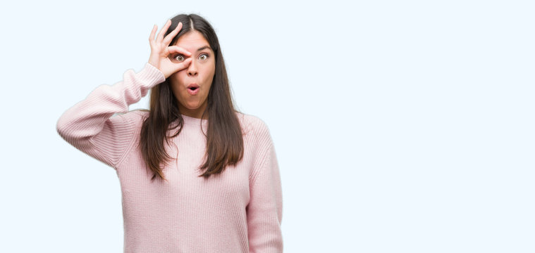 Young beautiful hispanic woman wearing a sweater doing ok gesture shocked with surprised face, eye looking through fingers. Unbelieving expression.
