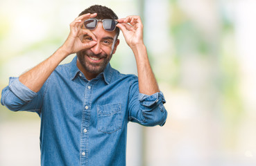 Naklejka premium Adult hispanic man wearing sunglasses over isolated background doing ok gesture with hand smiling, eye looking through fingers with happy face.