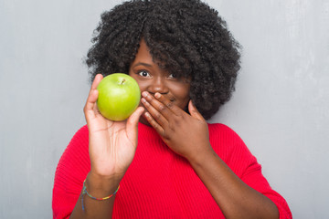 Young african american woman over grey grunge wall eating green apple cover mouth with hand shocked with shame for mistake, expression of fear, scared in silence, secret concept