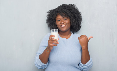 Young african american woman over grey grunge wall drinking a glass of milk pointing and showing with thumb up to the side with happy face smiling