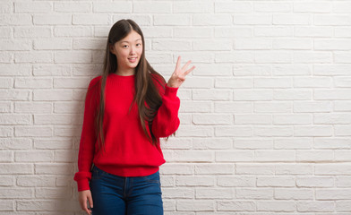 Obraz na płótnie Canvas Young Chinese woman over brick wall showing and pointing up with fingers number three while smiling confident and happy.