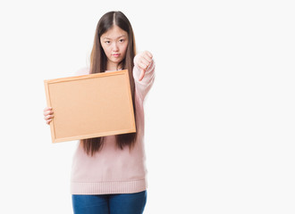 Young Chinese woman over isolated background holding corkboard with angry face, negative sign showing dislike with thumbs down, rejection concept