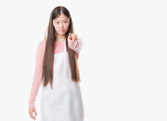 Young Chinese shop owner woman over isolated background wearing white apron pointing with finger to the camera and to you, hand sign, positive and confident gesture from the front