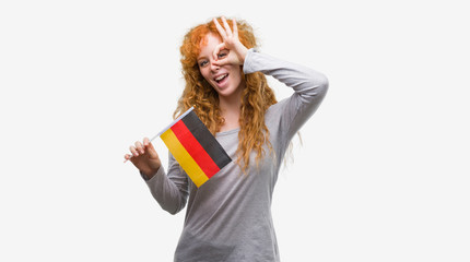Young redhead woman holding flag of Germany with happy face smiling doing ok sign with hand on eye looking through fingers