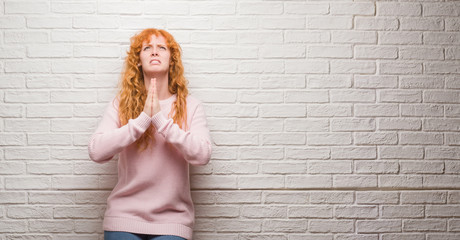 Young redhead woman standing over brick wall begging and praying with hands together with hope expression on face very emotional and worried. Asking for forgiveness. Religion concept.