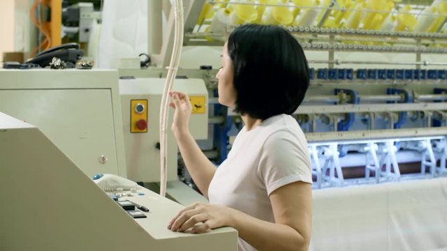 Female Asian technician pressing buttons on computer while turning on industrial textile machine at plant and then looking at camera and smiling