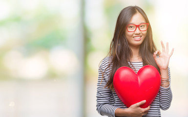 Young asian woman in love holding read heart over isolated background doing ok sign with fingers, excellent symbol