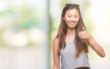 Young asian woman wearing sunglasses over isolated background doing happy thumbs up gesture with hand. Approving expression looking at the camera with showing success.