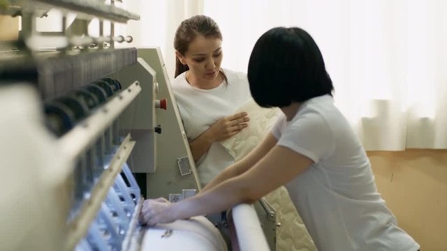 Tilt up of two young female colleagues working with textile machine in factory and discussing fabric