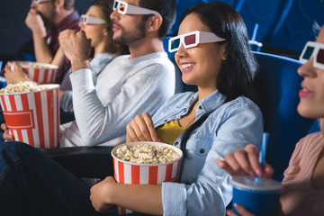 smiling multiethnic friends in 3d glasses with popcorn watching film together in movie theater