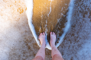 A young pale womens feet standing on the sand with a wave around her feet