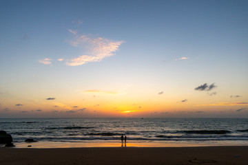 A landscape of a beach just after sunset with two people silhouette standing on the sand by, water in the background