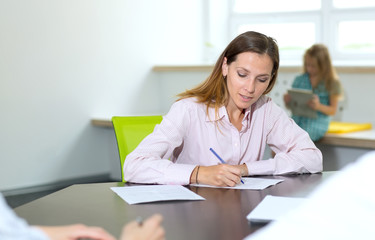 Woman signs documents sitting at a table on the background of her daughter. Mom makes out an application or questionnaire for the child's school.