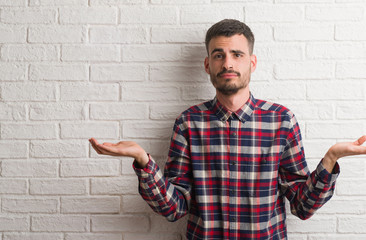Young adult man standing over white brick wall clueless and confused expression with arms and hands...