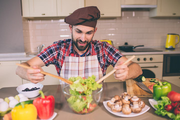 Careful and concentrated man stands at table and holds tow wooden spoons. Guy is mixing salad in glass bowl. THere are different vegetables on table.