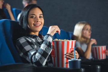 smiling asian woman with popcorn looking at camera in cinema