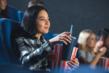 side view of smiling asian woman with popcorn and soda drink watching movie in cinema