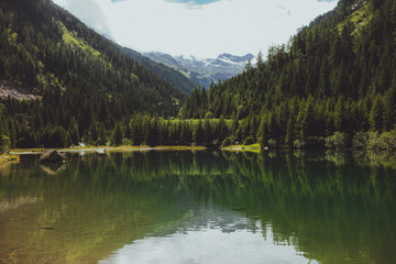 green lake with a reflection of clouds surrounded by mountains and tall pine trees
