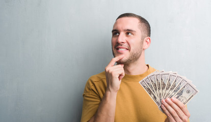 Young caucasian man over grey grunge wall holding dollars serious face thinking about question,...