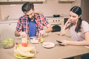 Happy guy sits and table and eats omlet. He cuts food into pieces with knife and fork. Girl looks at phone she holds in hands. She is bored and tired.