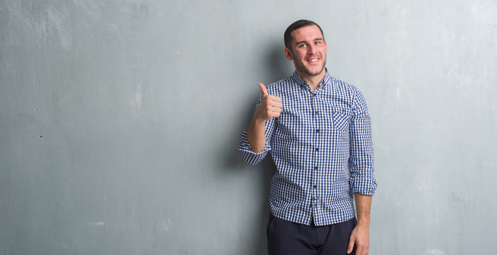 Young caucasian man over grey grunge wall doing happy thumbs up gesture with hand. Approving expression looking at the camera with showing success.