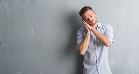 Young redhead business man over grey grunge wall sleeping tired dreaming and posing with hands together while smiling with closed eyes.