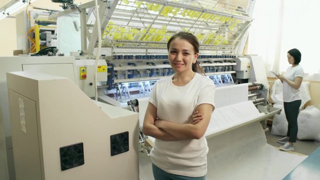 Young brunette woman standing in textile factory, smiling and looking at camera while her female colleague using digital tablet and operating textile machine