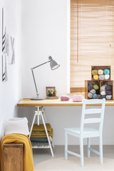 A simple, white chair and a gray desk lamp on a wooden surface in a white tailor workshop interior with colorful yarn and blankets. Real photo.
