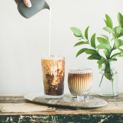 Iced coffee in tall glasses with milk poured over from pitcher by hand, white wall and green plant branches at background, copy space, square crop. Summer refreshing beverage ice coffee concept