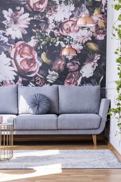 Grey sofa against flowers wallpaper in feminine living room interior with rug. Real photo