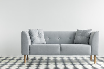 Grey sofa with pillows on patterned floor in minimal living room interior. Real photo